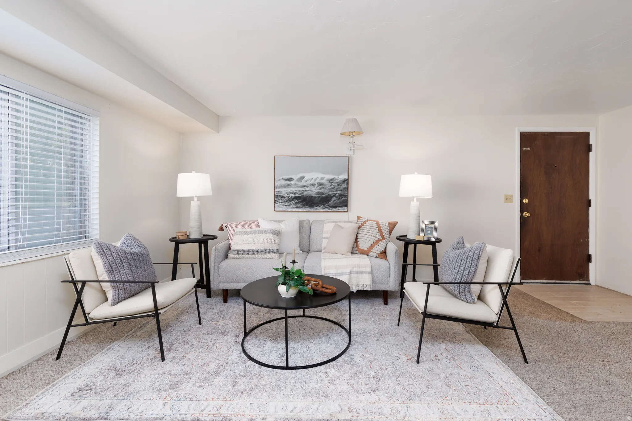 Bright living area featuring a large window with blinds, light-toned carpeting, and a contrasting dark wood-finish entry door