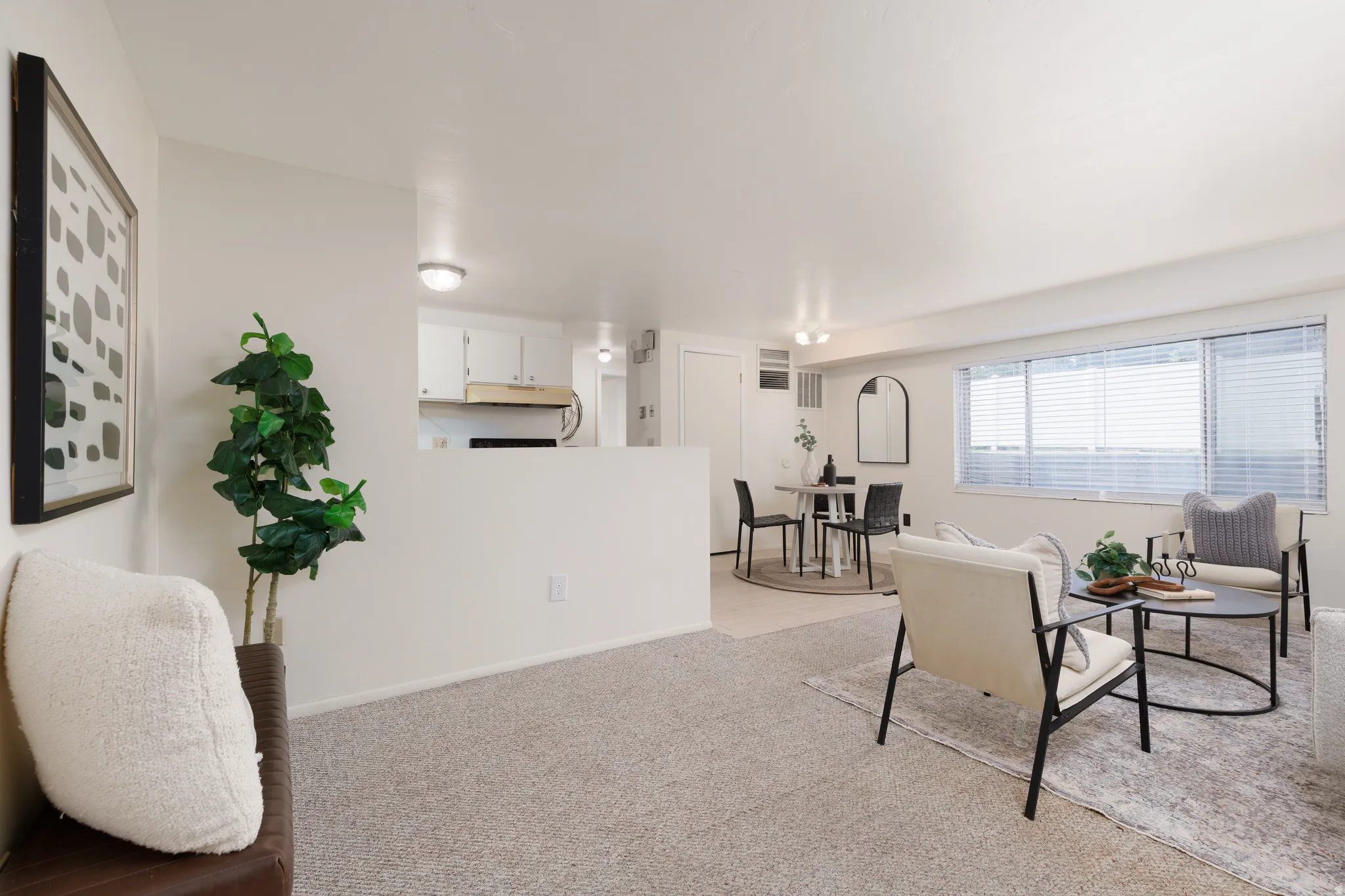 Open concept living space featuring neutral carpeting, a large picture window with horizontal blinds, and a dining area with a distinct flooring material