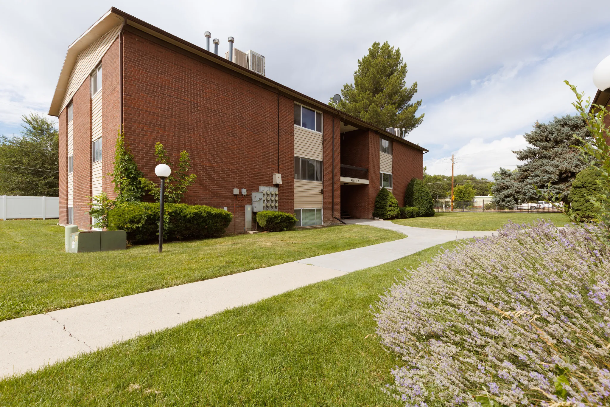 Brick exterior building with tan siding accents, featuring a concrete pathway, manicured lawn, established shrubs, and mature trees