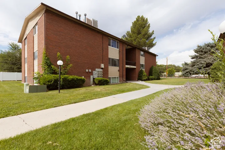 Brick exterior building with tan siding accents, featuring a concrete pathway, manicured lawn, established shrubs, and mature trees