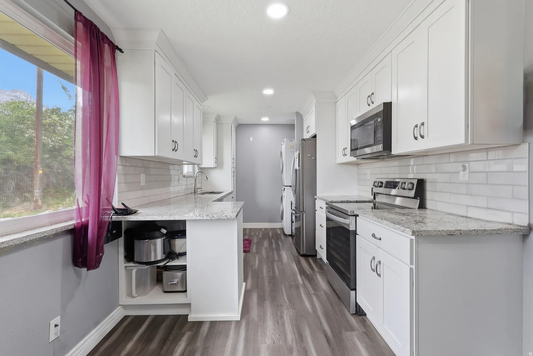 Kitchen featuring stainless steel appliances, light stone counters, white cabinets, stacked washer and dryer, and dark wood-style flooring