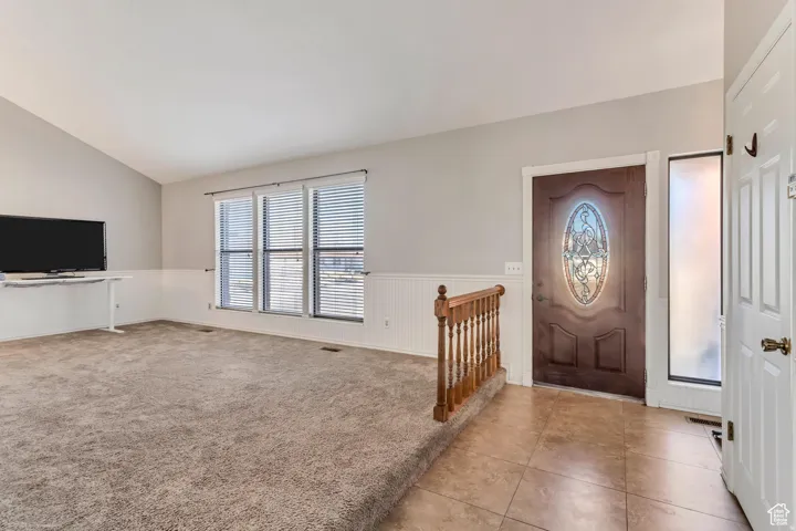 Vaulted ceiling, paneled accent wall, multiple windows with blinds, wood-finish railing, and tile flooring at entry