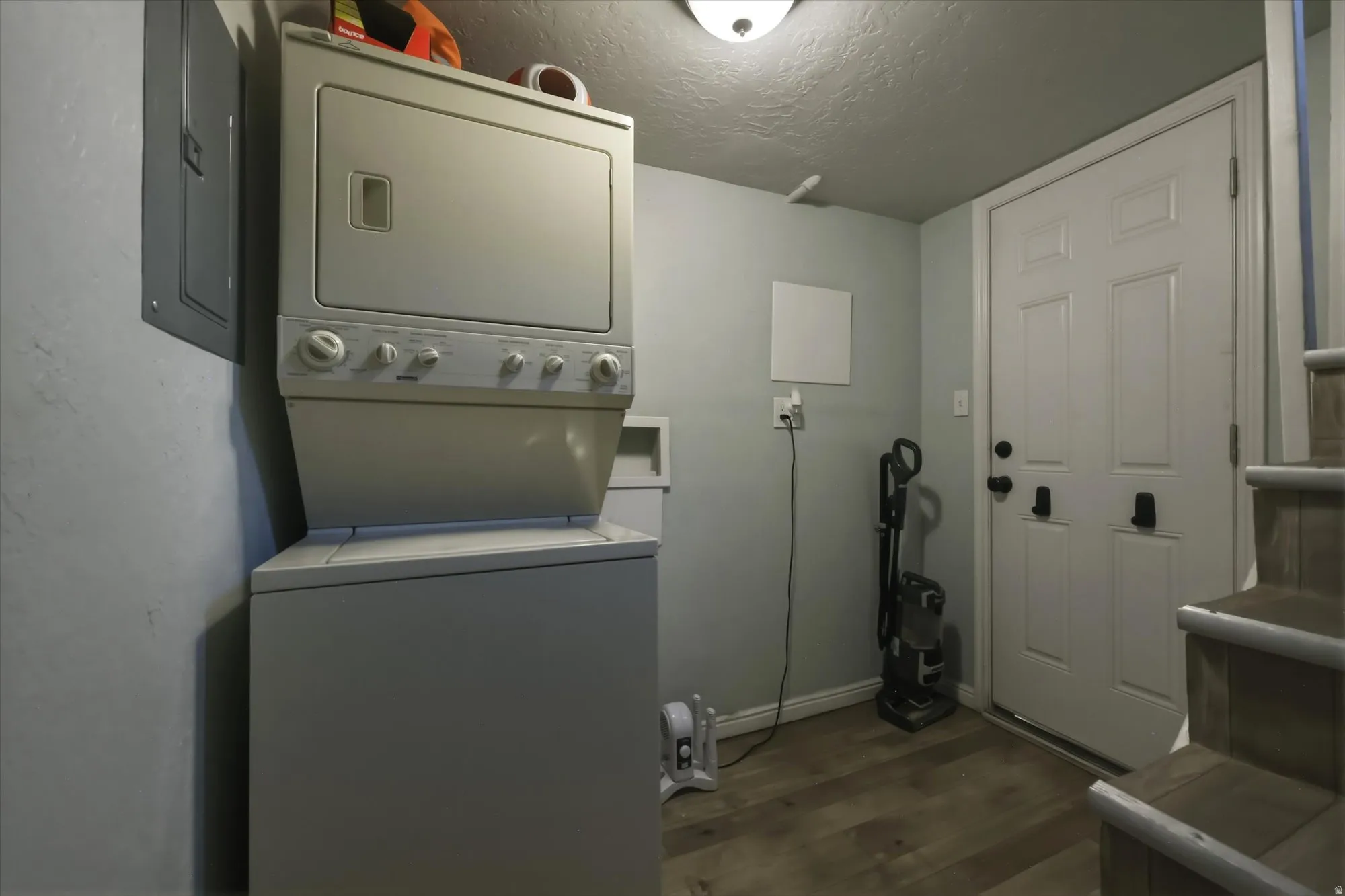 Laundry room with a textured ceiling, dark wood finished floors, stacked washer / dryer, and electric panel