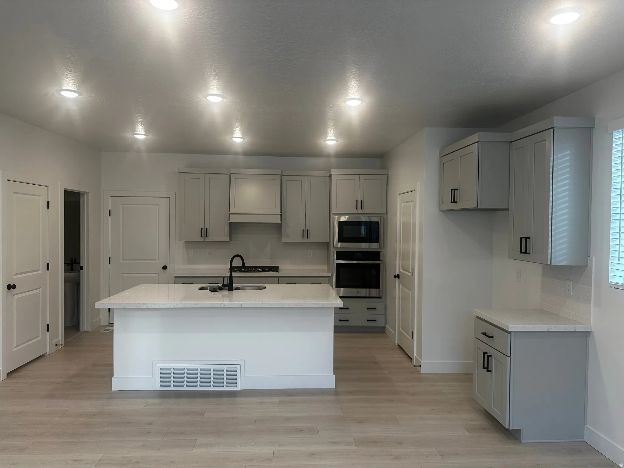 Kitchen with gray cabinetry, light wood-style flooring, stainless steel appliances, light stone countertops, and a kitchen island with sink