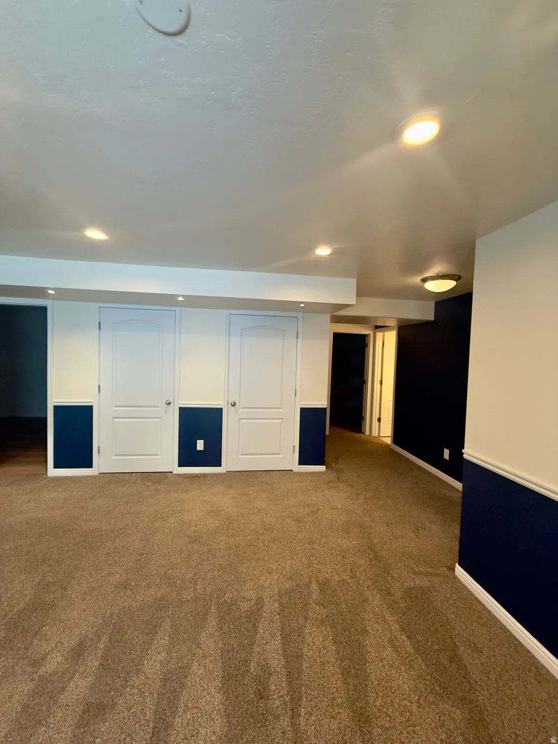 Spacious room featuring plush brown carpeting, recessed ceiling lighting, and white walls with a dark blue wainscoting accent