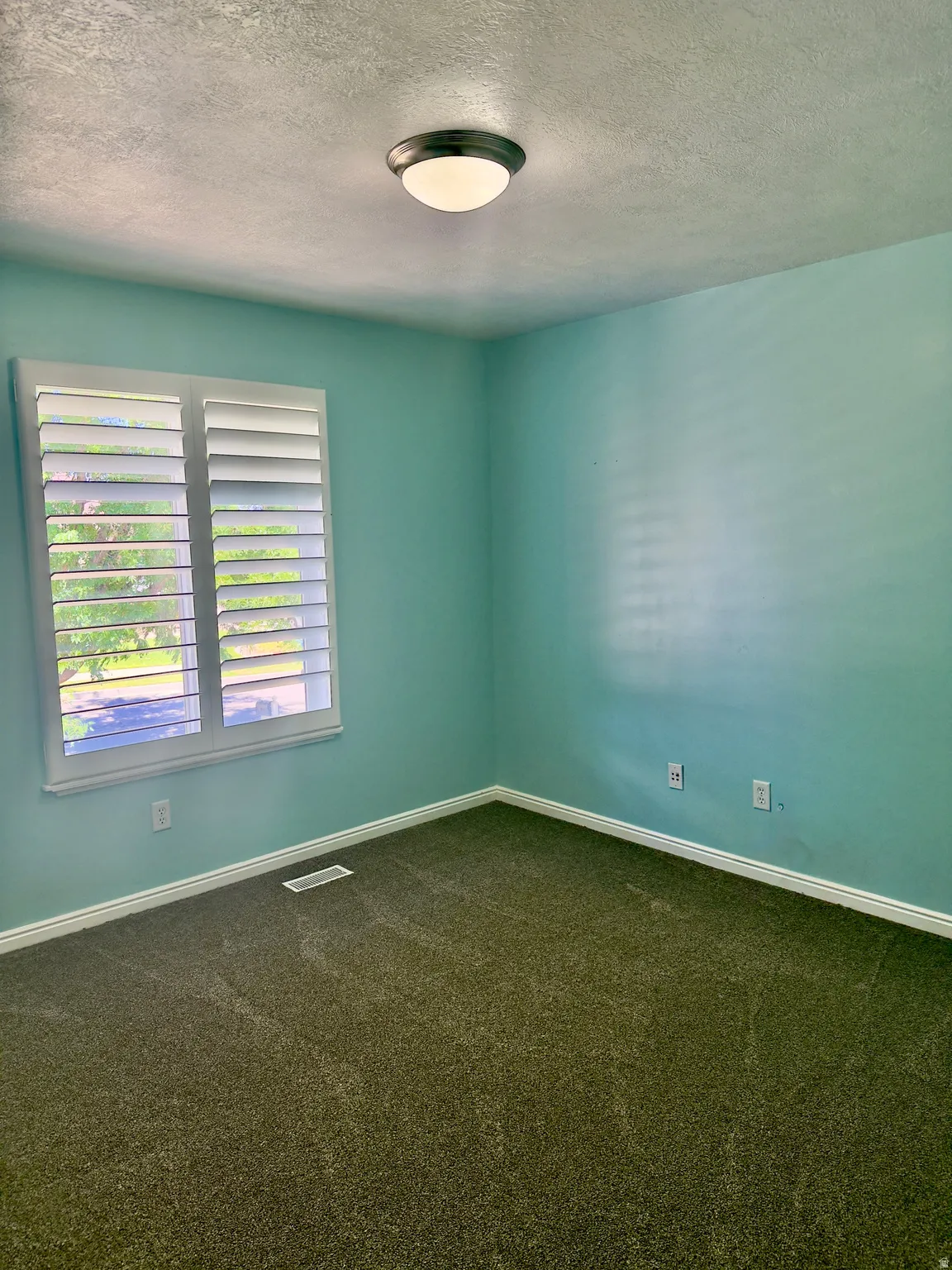 Room featuring a window with white louvered shutters, light blue walls, and dark carpet flooring