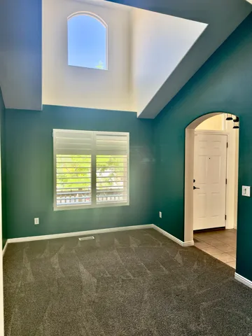 Room featuring high vaulted ceilings, an arched upper window, a rectangular window with white louvered shutters, and dark gray carpeting