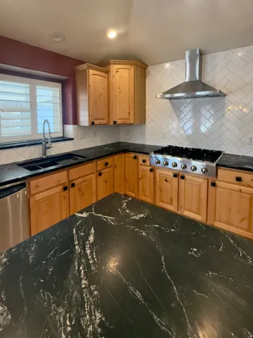 Kitchen featuring natural wood cabinetry, dark patterned countertops, a stainless steel gas range with a matching range hood, and a white herringbone tile backsplash