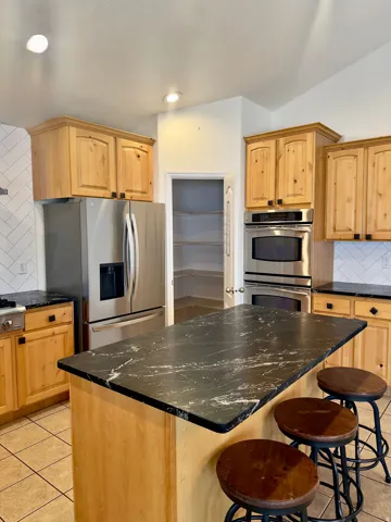 Kitchen featuring light wood cabinetry, stainless steel appliances, a center island with a dark stone countertop, subway tile backsplash, and ceramic tile flooring