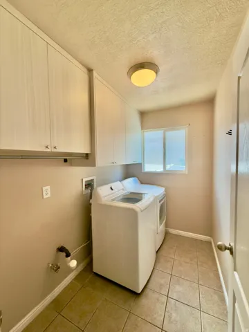 Dedicated laundry area featuring ceramic tile flooring, overhead cabinetry, a ceiling-mounted light fixture, and a window providing natural illumination