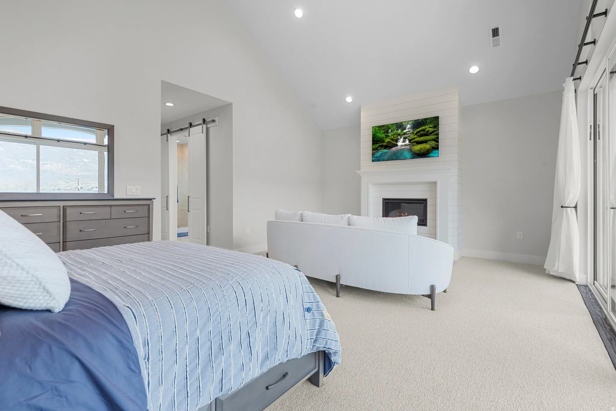 Bedroom with a barn door, vaulted ceiling, light carpet, ensuite bathroom, and a large fireplace