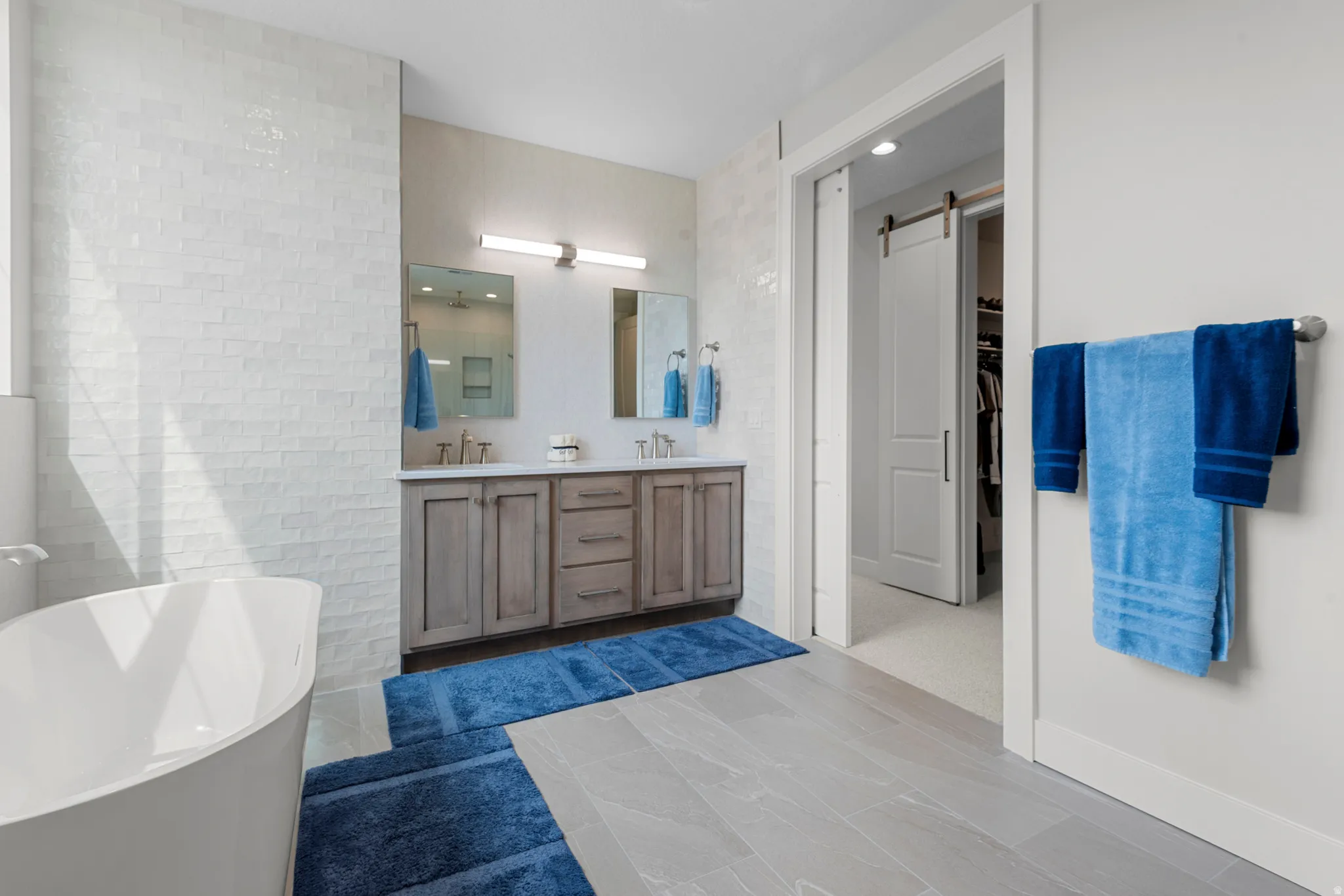 Bathroom with a freestanding bath, double vanity, tile walls, dark tile patterned floors, and a walk in closet
