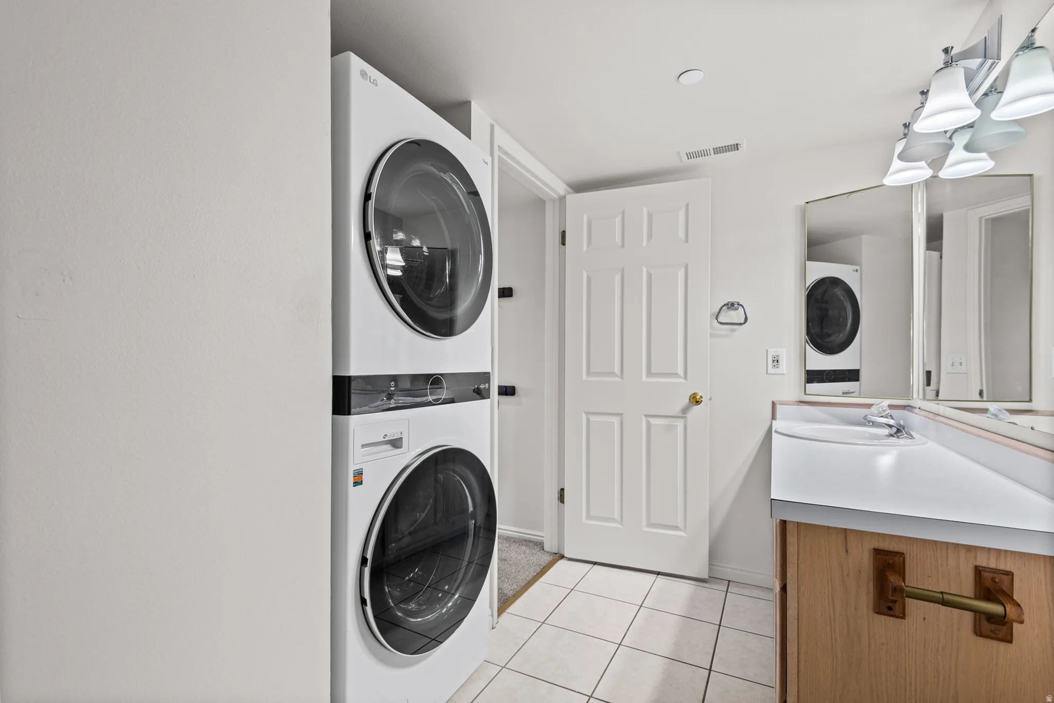 Stacked laundry appliances, white tiled flooring, single vanity with integrated sink, wall-mounted mirror, and a multi-bulb light fixture