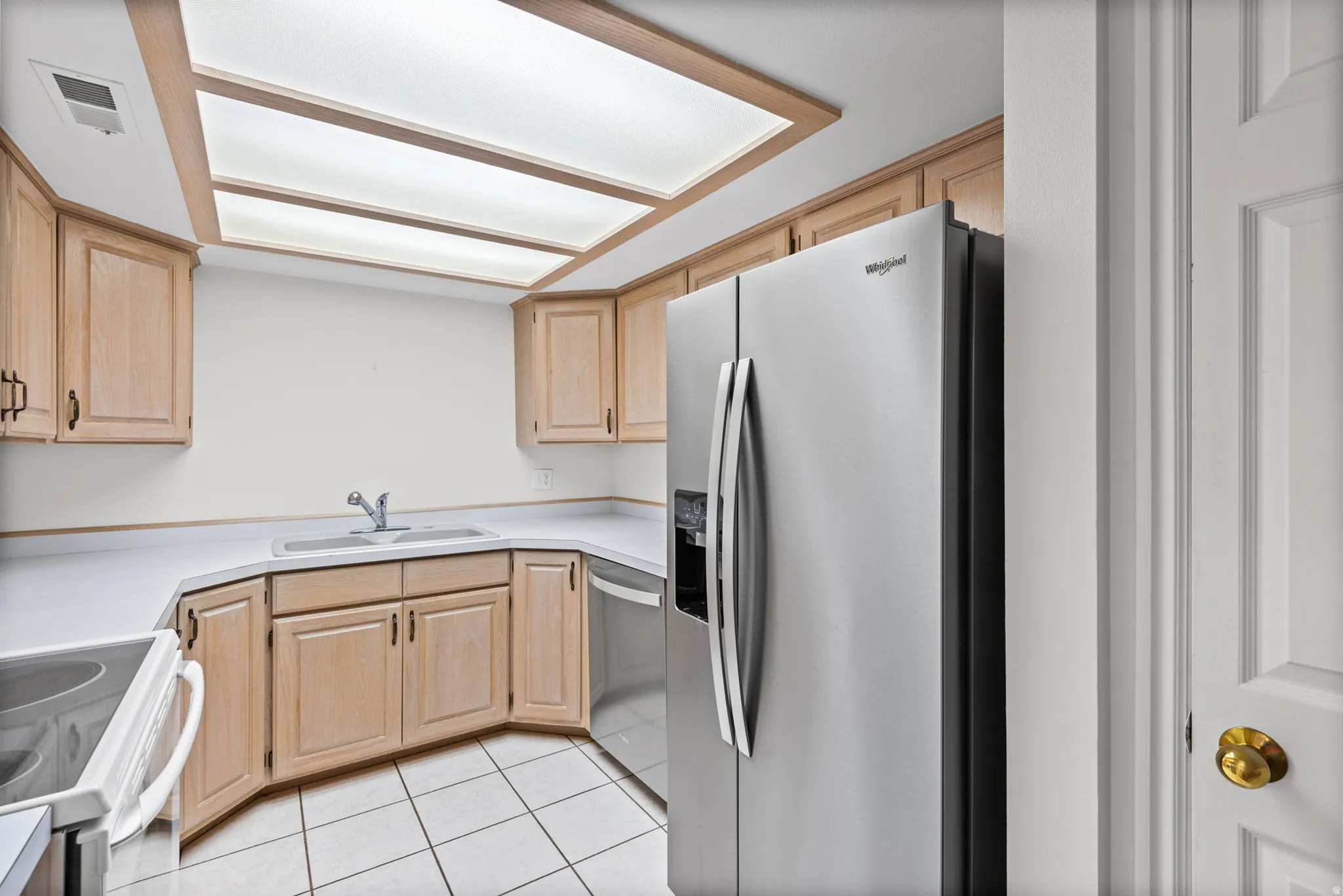 Neutral-toned cabinetry, white countertops, recessed ceiling light panels, stainless steel refrigerator, and tiled flooring