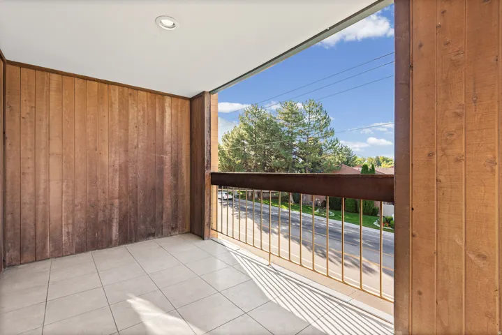 Spacious covered balcony with tile flooring, wood-finish paneled walls, and metal railing overlooking landscaped grounds and mature trees