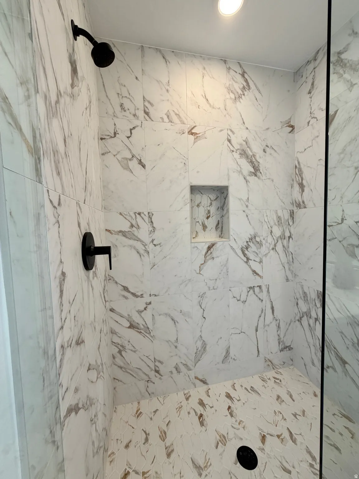 Shower enclosure featuring large format white marble-look wall tiles, matte black shower fixtures, a recessed niche, and a mosaic tile floor