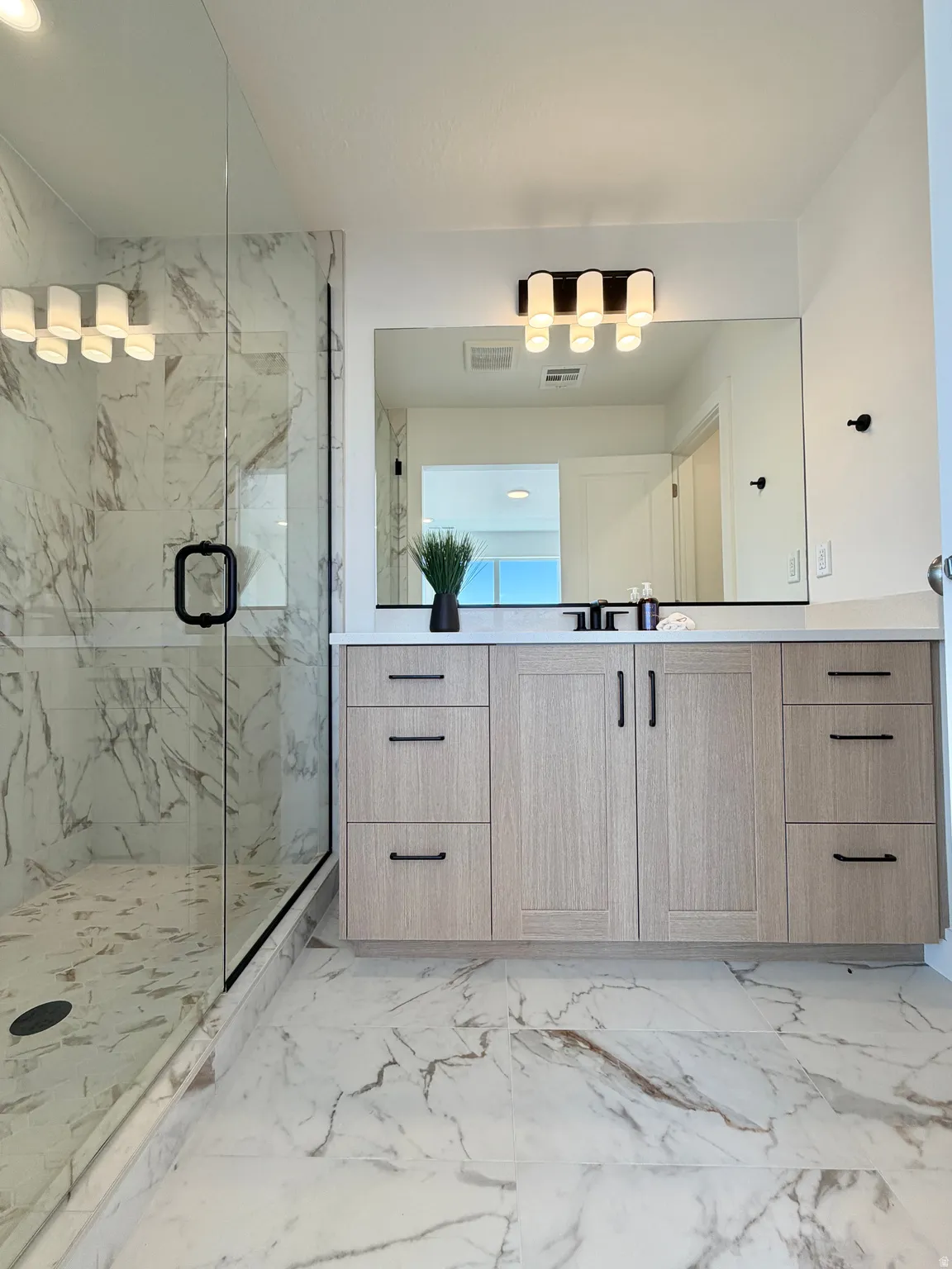 Contemporary bathroom featuring a spacious glass-enclosed shower with white marble-look tile, a light wood-finish vanity with black hardware, a rectangular vanity mirror, and a modern five-bulb light fixture