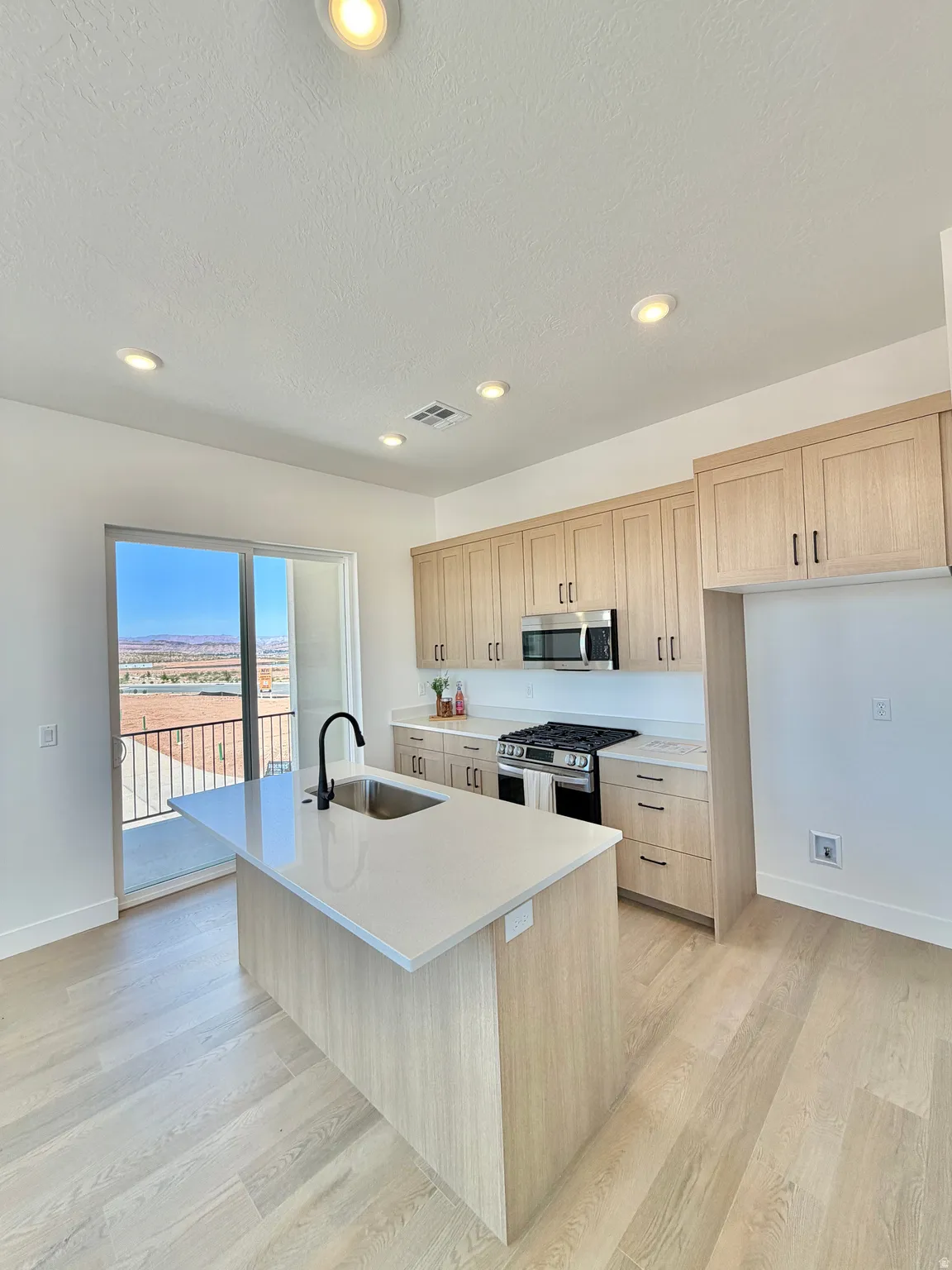 Modern kitchen featuring a central island with an undermount sink and black faucet, light wood-finish cabinetry, stainless steel appliances, and light-toned flooring
