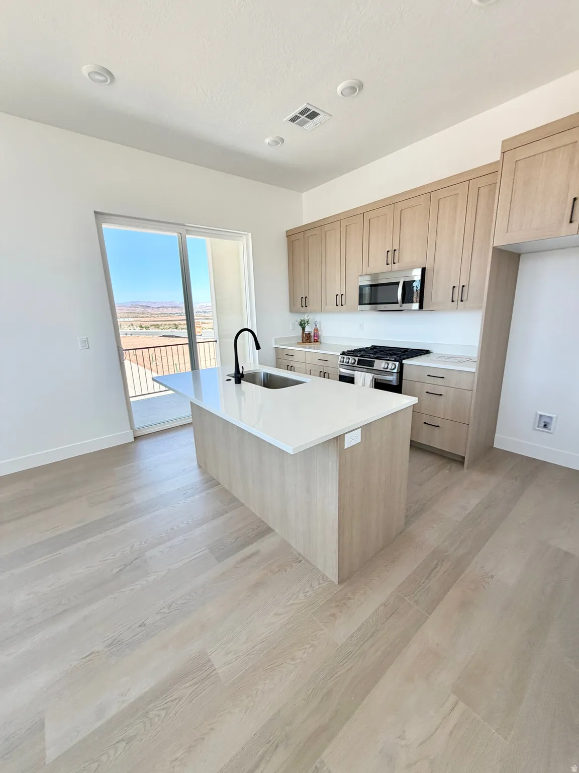 Modern kitchen featuring light wood-finish cabinetry, stainless steel appliances, and a central island with a sink and white countertop