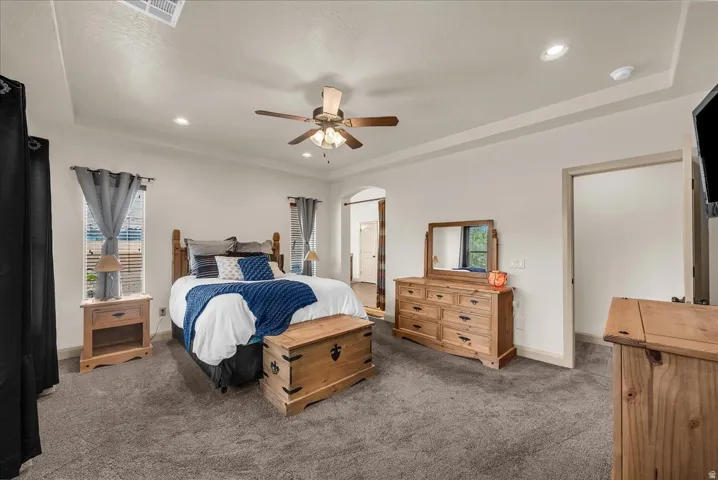 Primary bedroom with dark colored carpet, multiple windows, ceiling fan, arched walkways, and recessed lighting