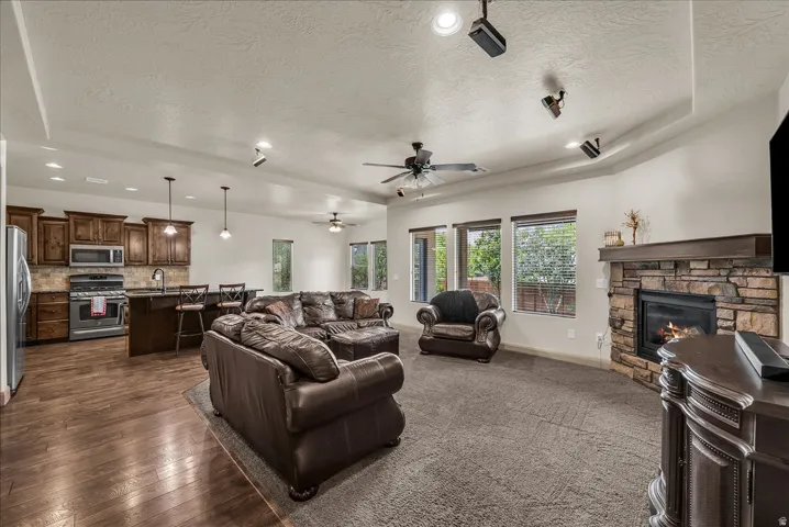 Living room featuring a raised ceiling, a textured ceiling, a stone fireplace, a ceiling fan, and dark wood finished floors