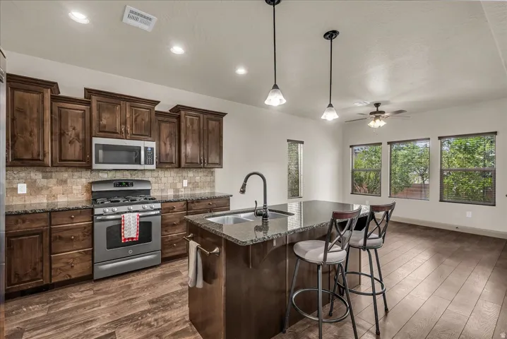 Kitchen featuring stainless steel appliances, dark wood finish cabinetry, an island with sink, backsplash, and dark stone counters