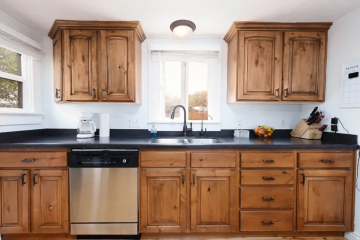 Kitchen featuring dark countertops, stainless steel dishwasher, wood finish cabinetry, and healthy amount of natural light