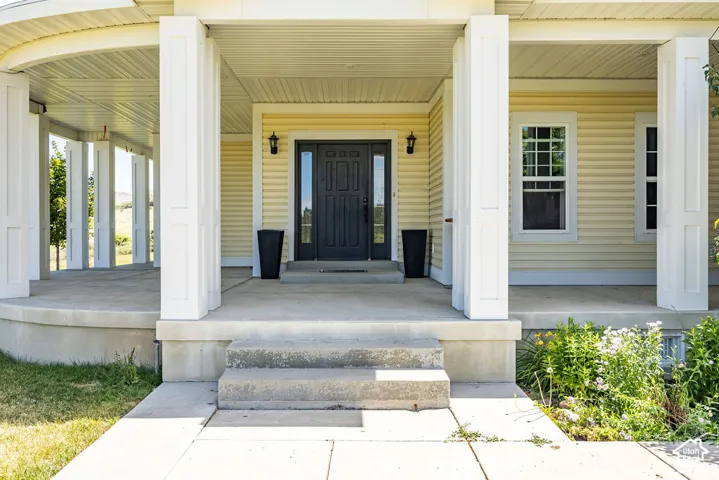 Inviting wrap-around front porch featuring a dark entry door with sidelights, a light yellow siding exterior, white trim, and multiple double-hung windows