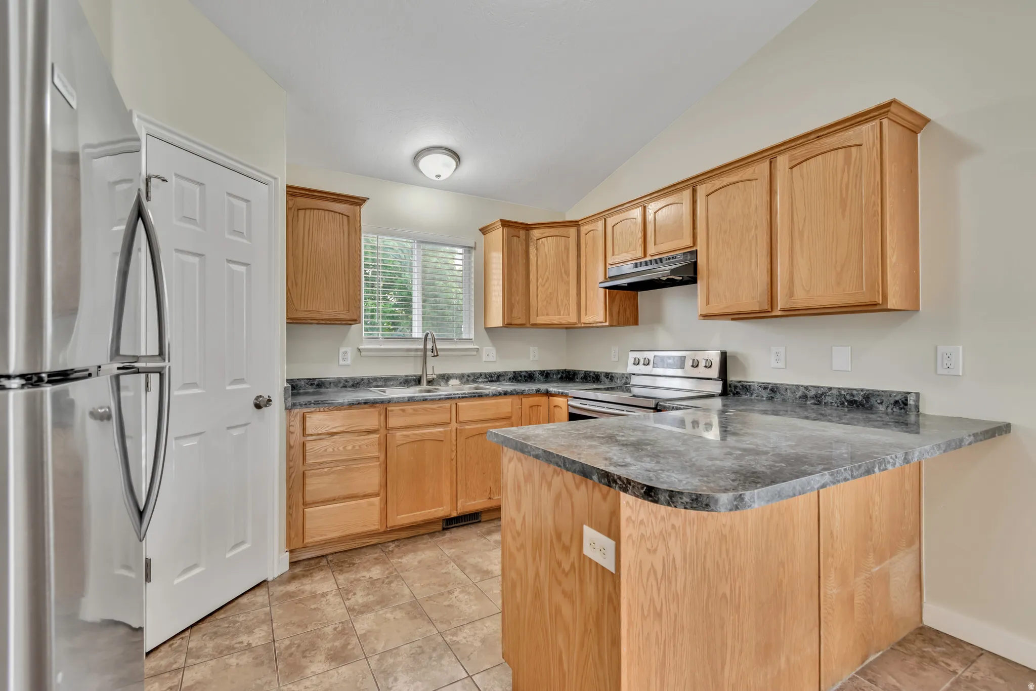 Kitchen featuring stainless steel appliances, a peninsula, vaulted ceiling, dark countertops, and light tile patterned floors