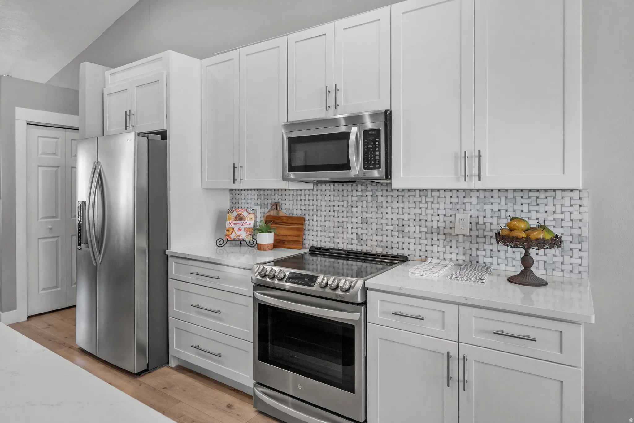 Kitchen featuring stainless steel appliances, white cabinets, quartz countertops, light wood LVP flooring, and tile backsplash