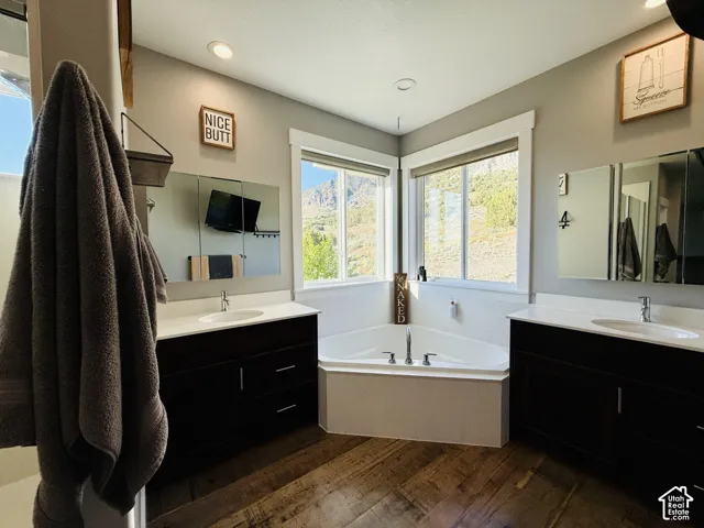 Corner soaking tub with surrounding windows, dual dark wood-finish vanities with white countertops, and wood-finish flooring