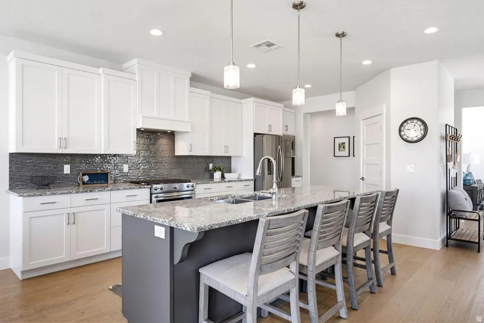 Two tone kitchen with light stone counters, light wood-style flooring, a center island with sink, stainless steel appliances, and decorative light fixtures