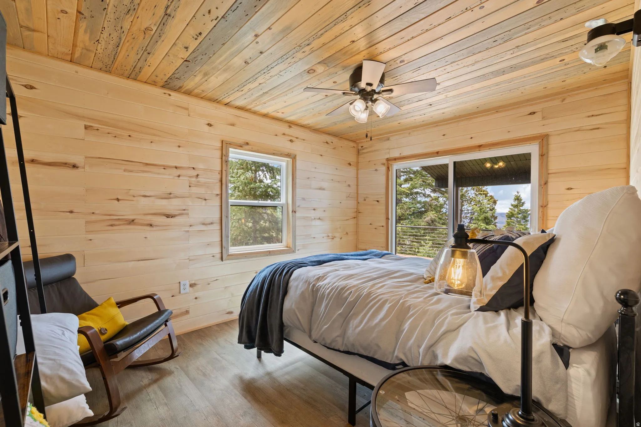 Cabin 1 - West; Bedroom with wood walls, wooden ceiling, wood finished floors, and a ceiling fan. Sliding door exiting onto the oversized deck