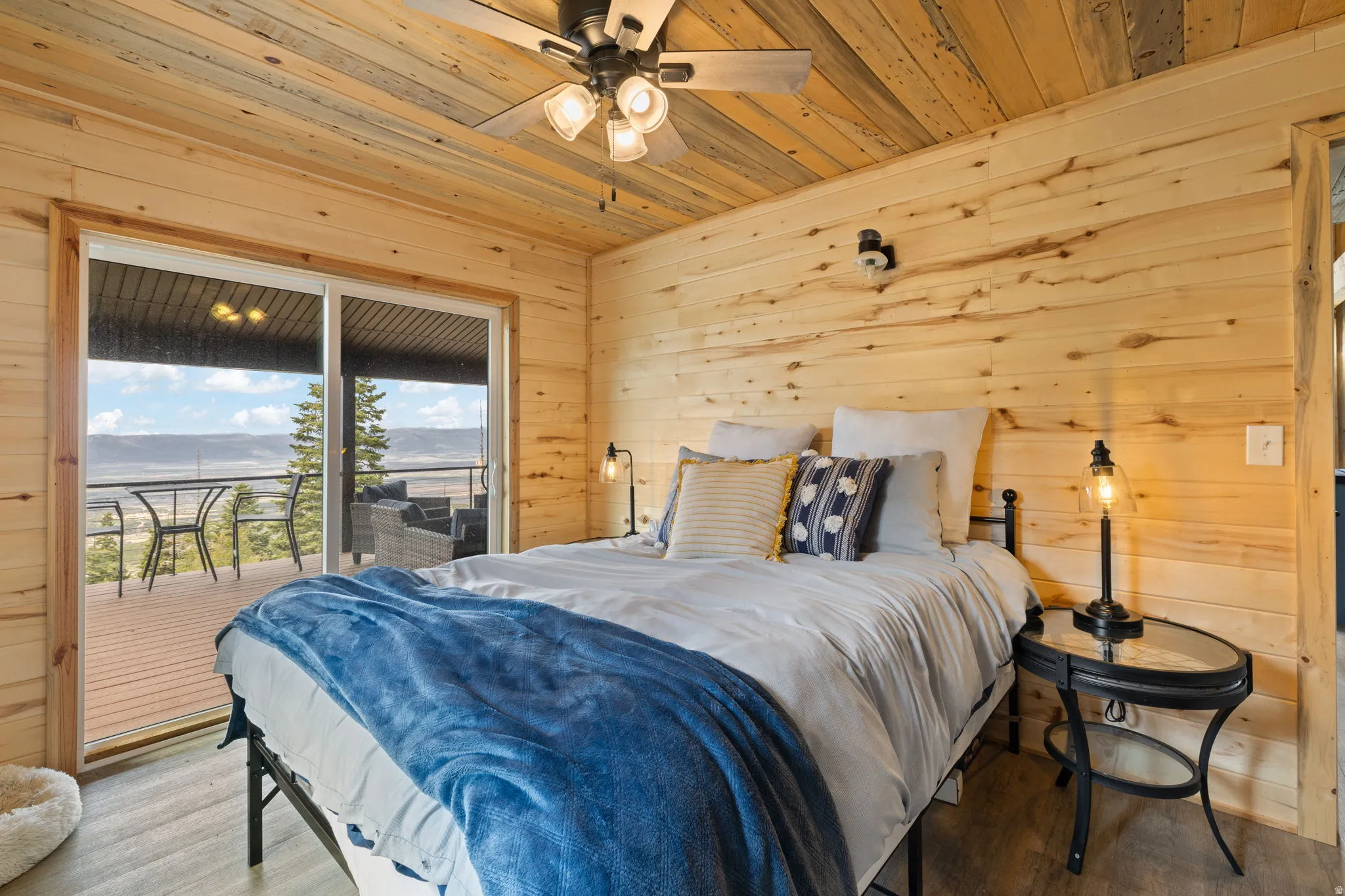 Cabin 1 - West; Bedroom featuring access to outside, wooden walls, wood ceiling, wood finished floors, and a ceiling fan. Sliding door exiting onto the oversized deck
