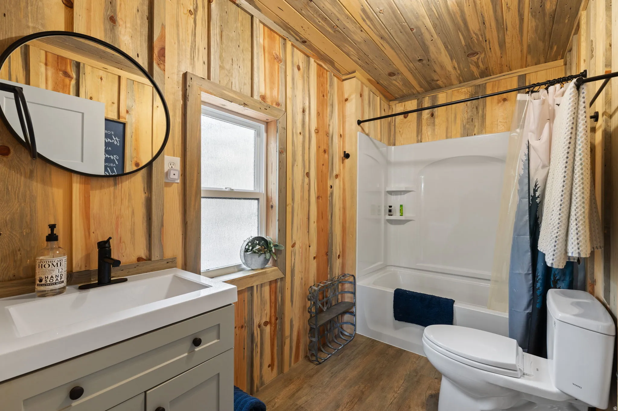 Cabin 1 - West; Full bath featuring wooden walls, vanity, dark wood finished floors, shower / bathtub combination with curtain, and wood ceiling