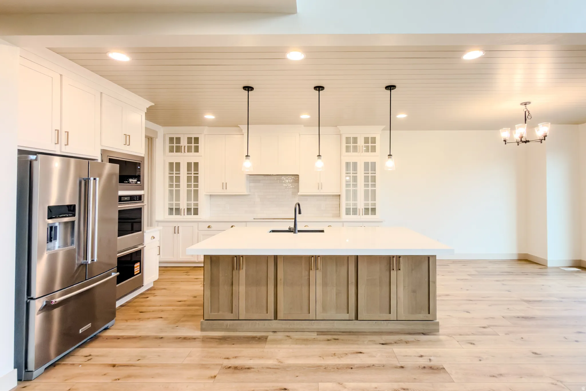 Two tone kitchen with glass fronted cabinets, stainless steel appliances, an island with sink, light wood-style flooring, and suspended lighting