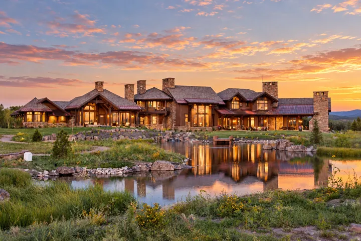 Expansive property featuring a stone and wood exterior, multiple stone chimneys, a private pond with a small dock, and a mountainous backdrop