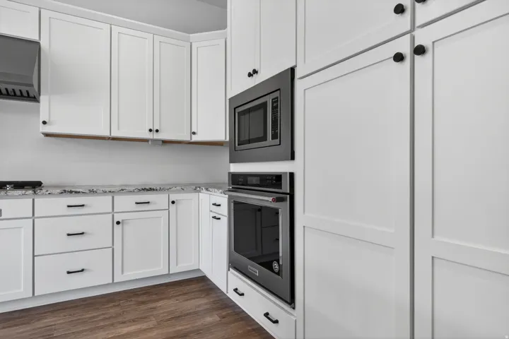 Kitchen featuring white shaker-style cabinetry, dark hardware, light countertops with grey veining, built-in microwave and oven, and wood-finish flooring