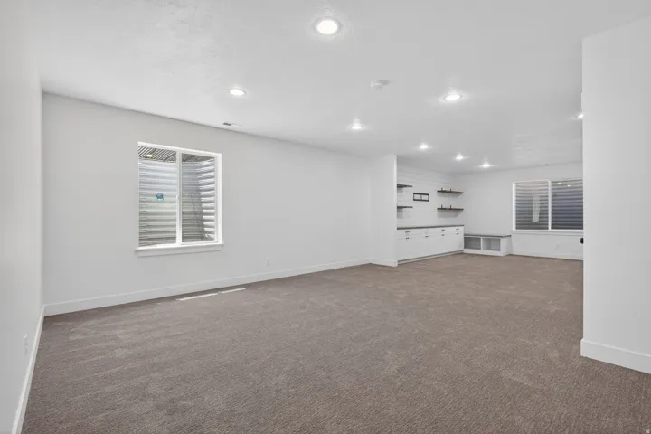 Spacious room featuring neutral carpeting, recessed lighting, two windows with horizontal blinds, built-in cabinetry, and floating shelves
