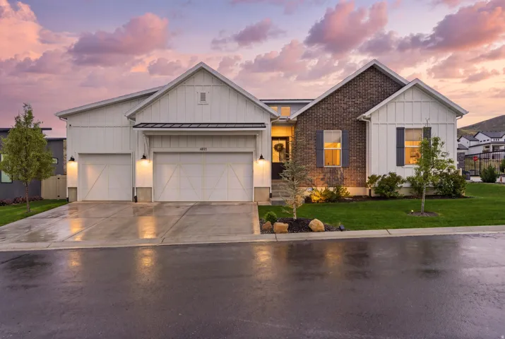 Modern farmhouse exterior featuring vertical white siding, dark brick accents, a two-car garage with carriage-style doors, a concrete driveway, and a manicured lawn