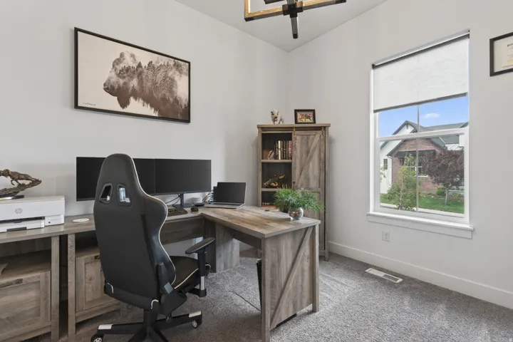 Dedicated workspace featuring a large window with roller shade, neutral wall paint, and gray carpet flooring