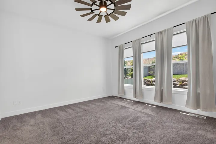 Spacious room featuring three large windows, a ceiling fan with unique wood-finish blades, and dark gray carpeting