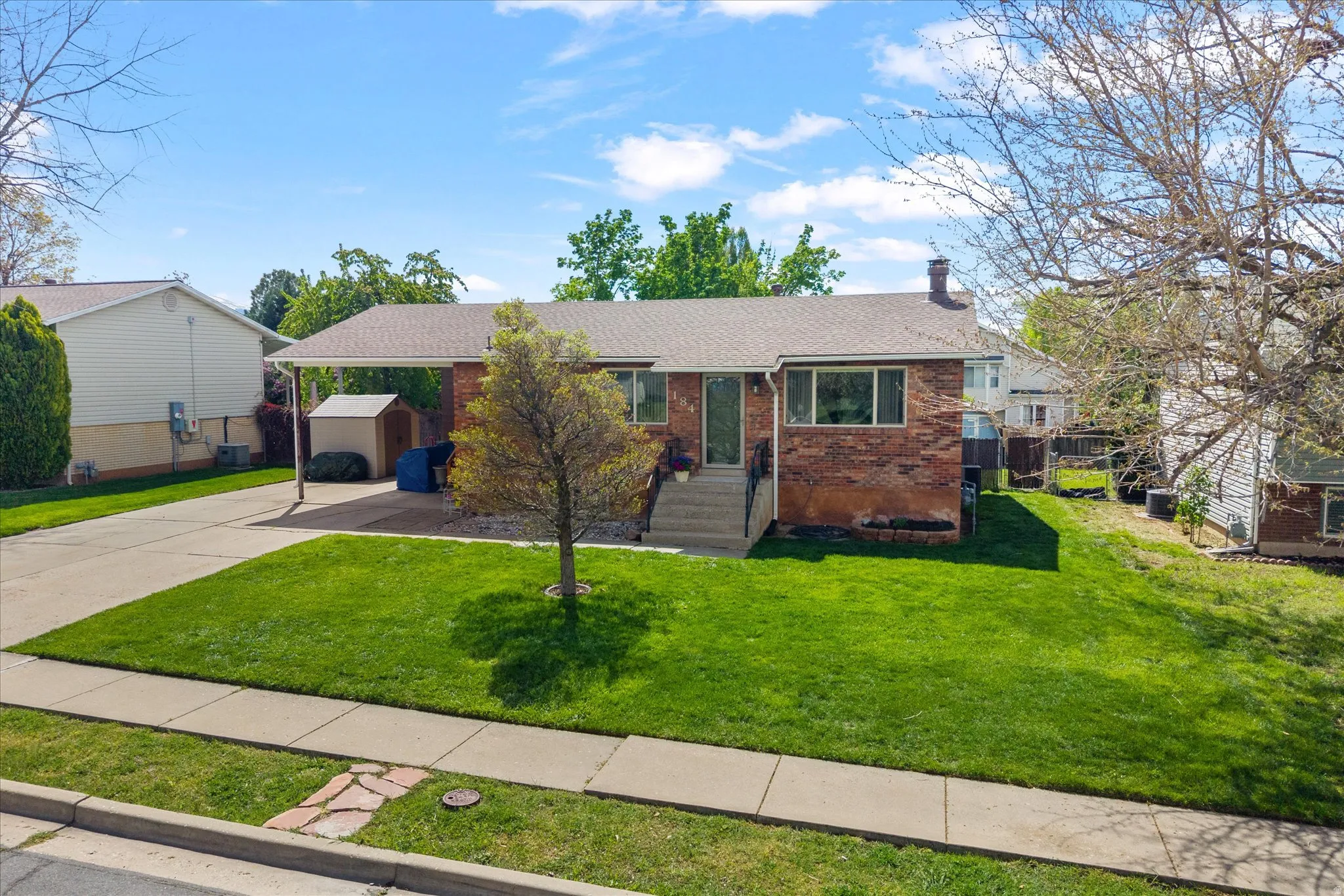 Single story home with a front yard, an attached carport, driveway, an outbuilding, and brick siding