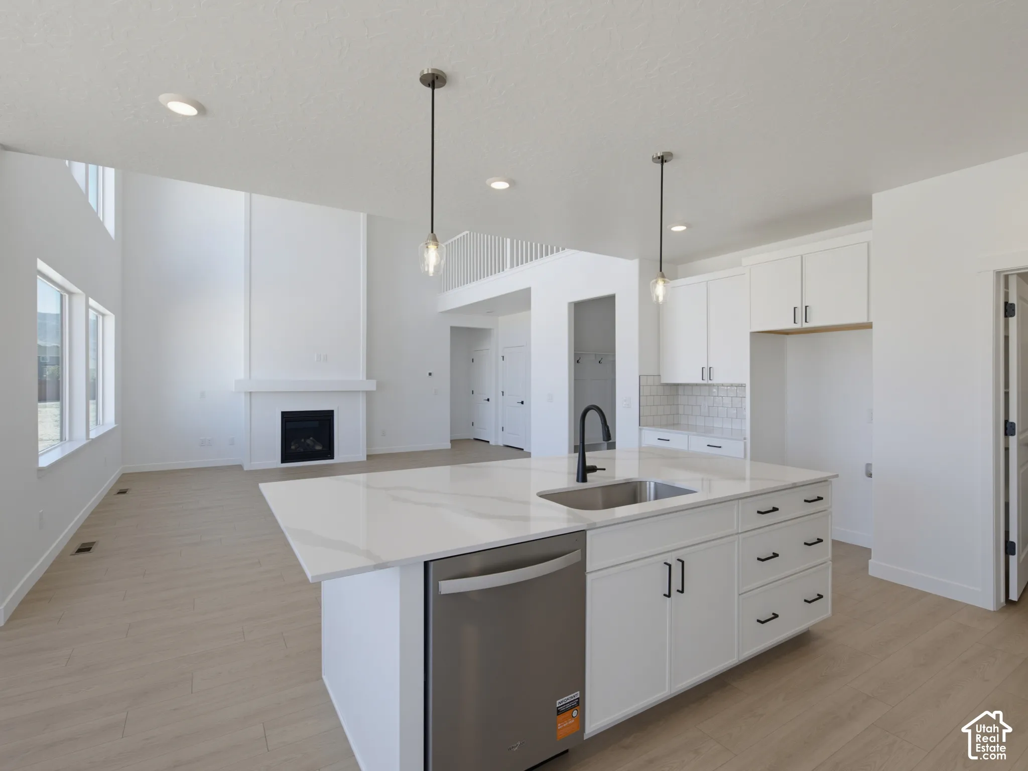 Open-concept main living area with high ceilings, featuring a gas fireplace with mantel, wood-finish flooring, and a kitchen island with sink and stainless steel dishwasher