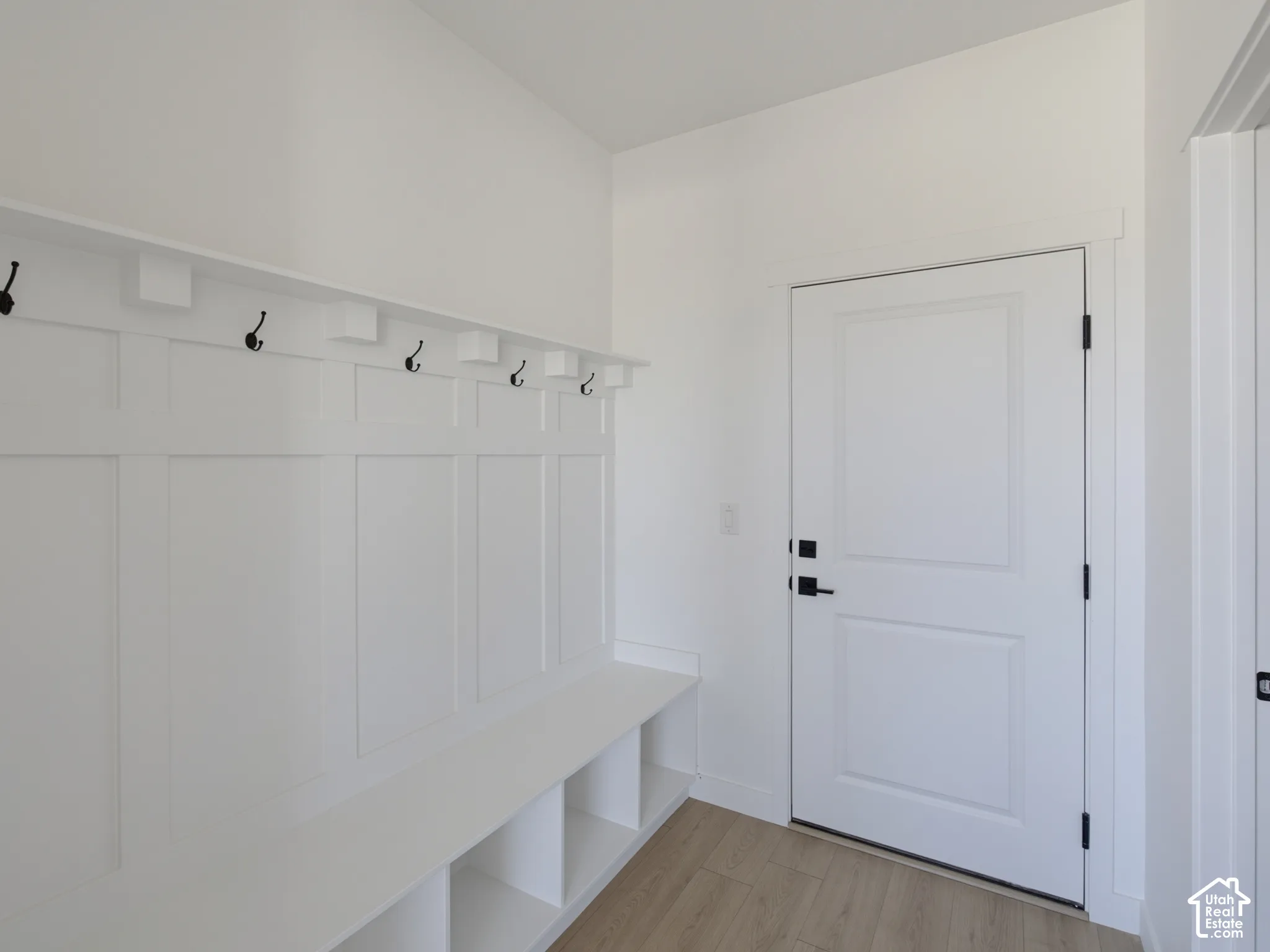 Built-in bench with integrated shelving, wainscoting paneling, multiple coat hooks, white paneled door, and wood-finish flooring