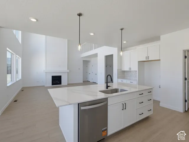 Open-concept main living area with high ceilings, featuring a gas fireplace with mantel, wood-finish flooring, and a kitchen island with sink and stainless steel dishwasher