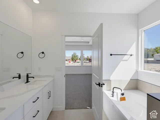 Dual vanity with integrated sinks and black fixtures, deep soaking tub with tile surround, large windows, recessed lighting, and white cabinetry with black hardware