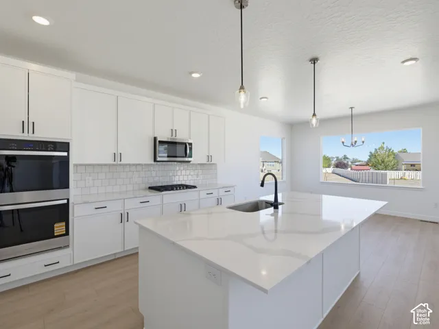 White cabinetry with dark hardware, a subway tile backsplash, built-in double ovens, a center island with an undermount sink, and wood-finish flooring