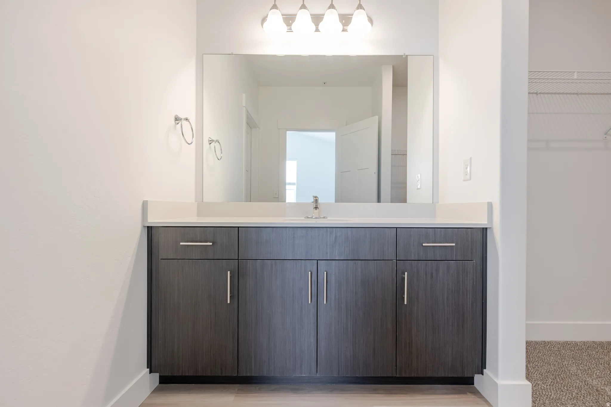 Wood-finish flooring, dark wood-finish vanity with chrome hardware, white countertop with integrated sink, rectangular mirror, and wall-mounted light fixture