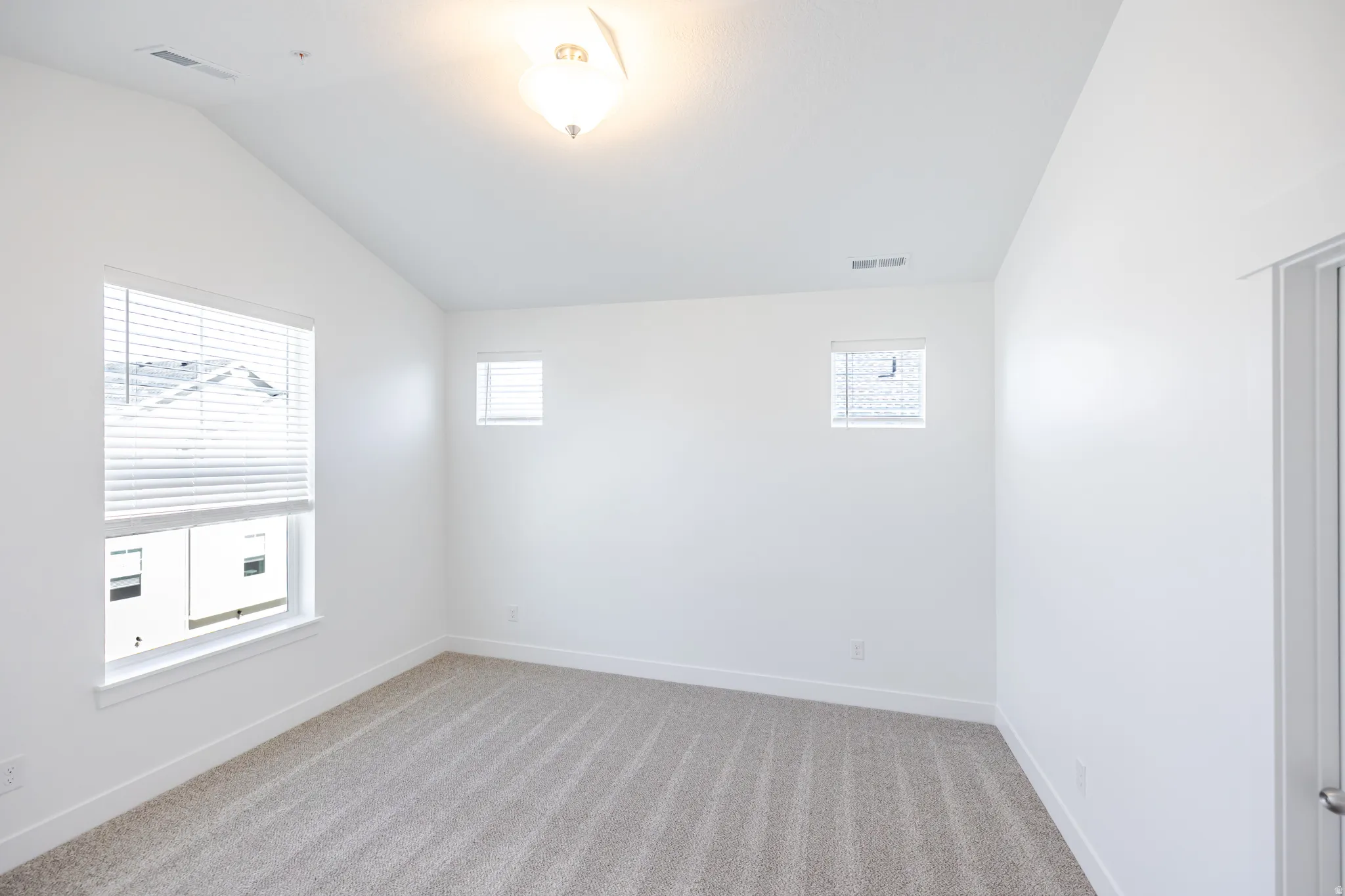 Vaulted ceiling with an overhead light fixture, multiple windows with blinds, and neutral carpeting